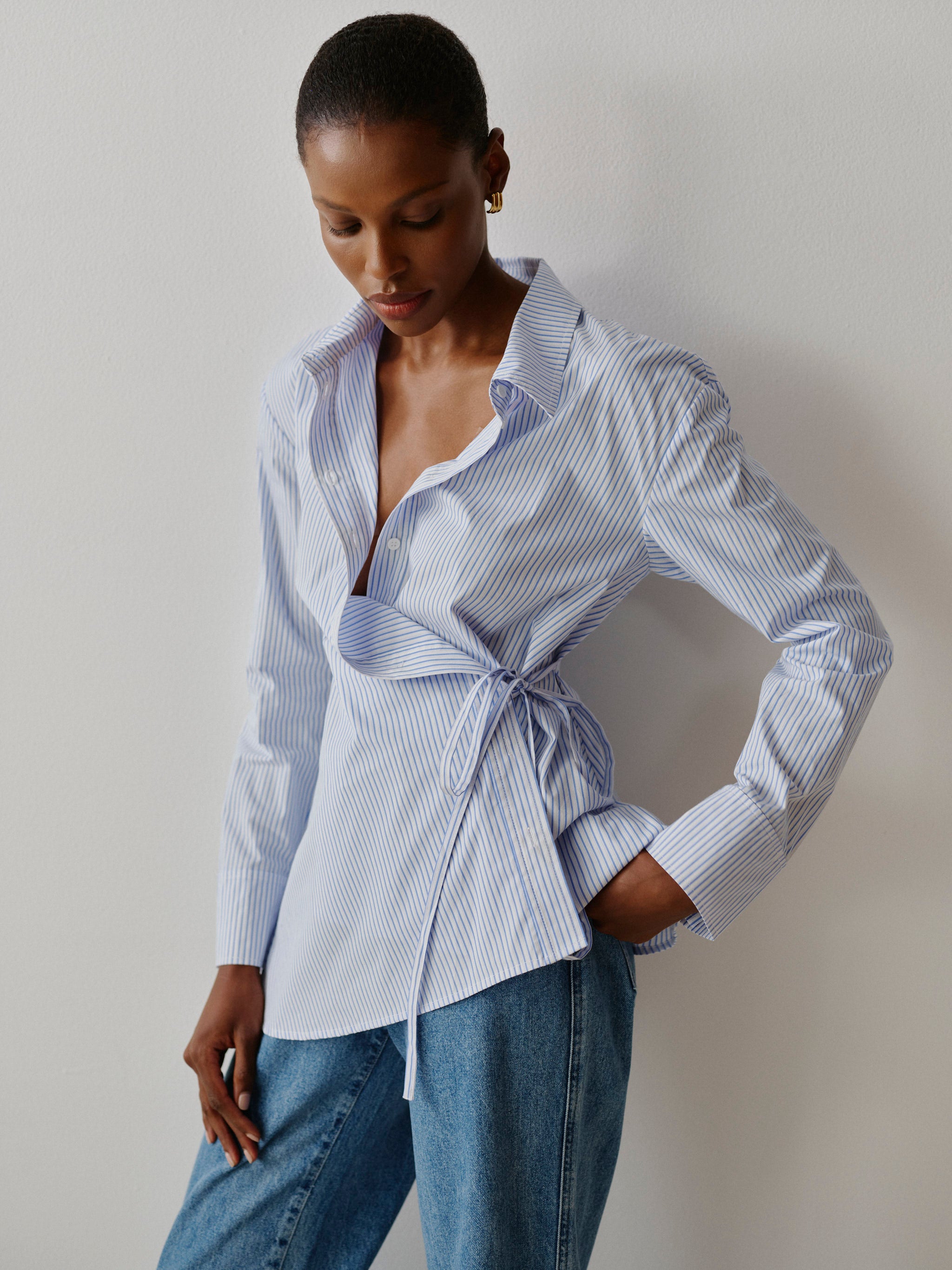 Woman wearing a blue and white striped shirt with a tie waist and jeans against a plain background