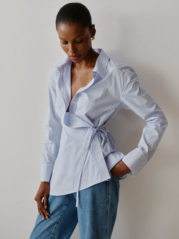 Woman wearing a blue and white striped shirt with a tie waist and jeans against a plain background
