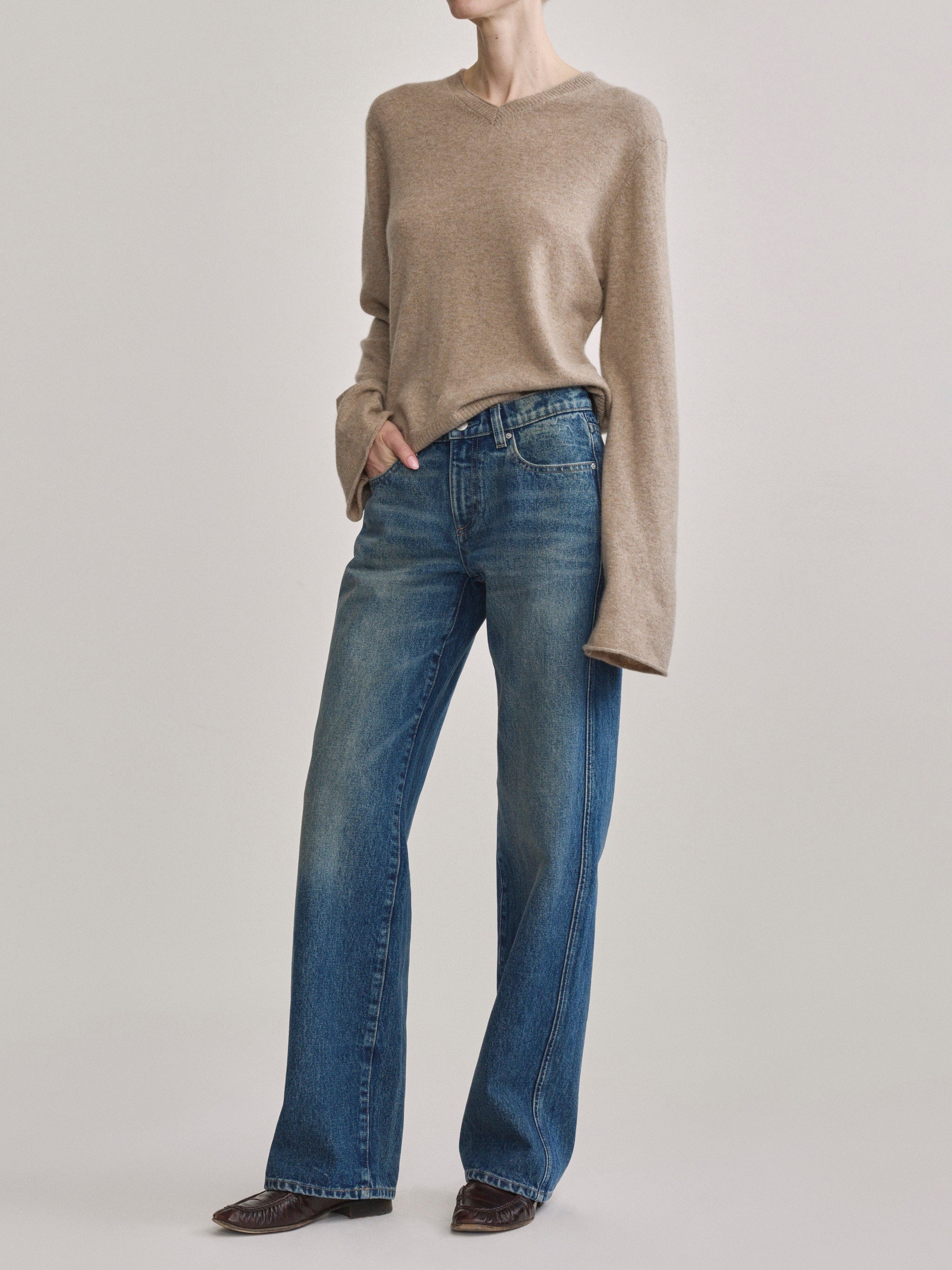 A model wearing a beige long-sleeve knit sweater and blue wide-leg jeans, standing against a light gray studio background.