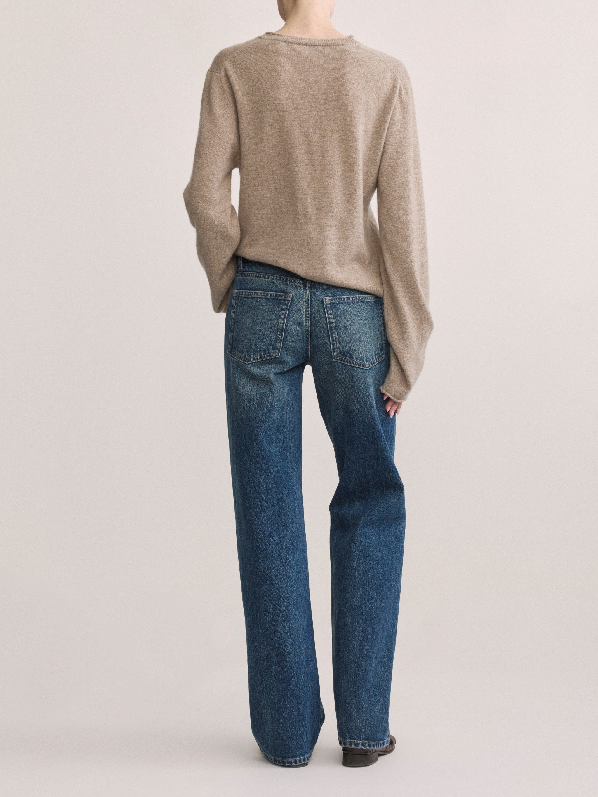 A model shown from the back wearing a beige knit sweater and blue wide-leg jeans against a light gray studio background.