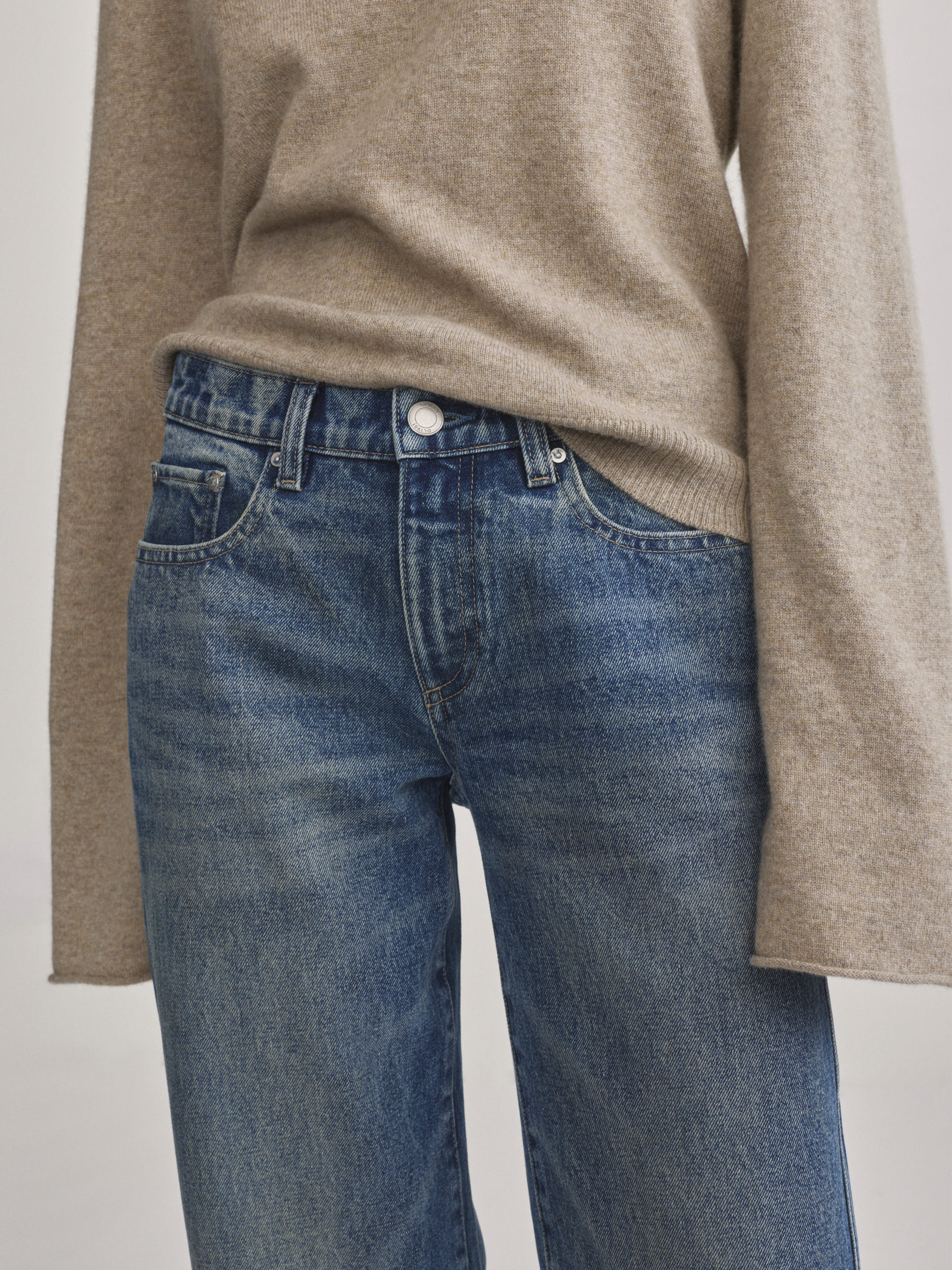 A close-up of a person wearing blue denim jeans and a beige knit sweater, cropped from the chest down against a plain light background.